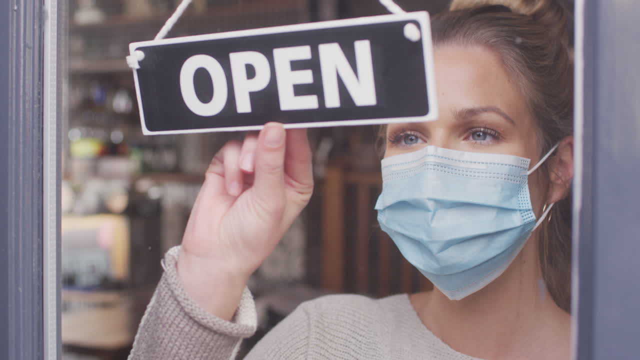 Proprietario femminile di una piccola impresa che indossa una maschera facciale che gira intorno al segno aperto durante la pandemia di salute