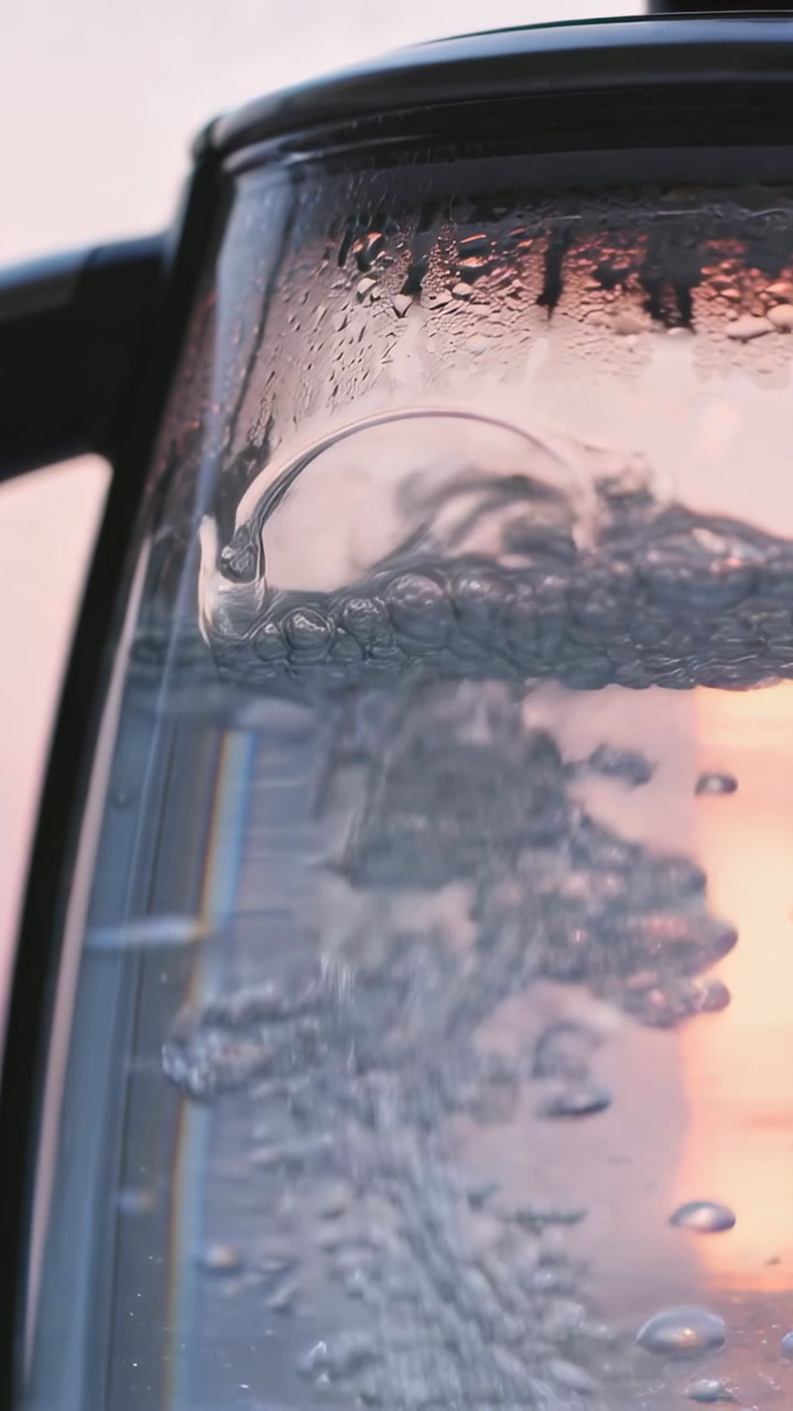 Vertical video: Heating glass kettle bubbling on counter beside window, left handle visible