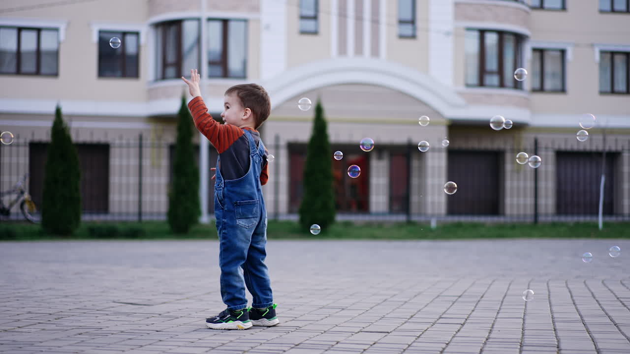 Caucasian toddler standing outdoors catches the soap bubbles. Active kid plays happily in the street.