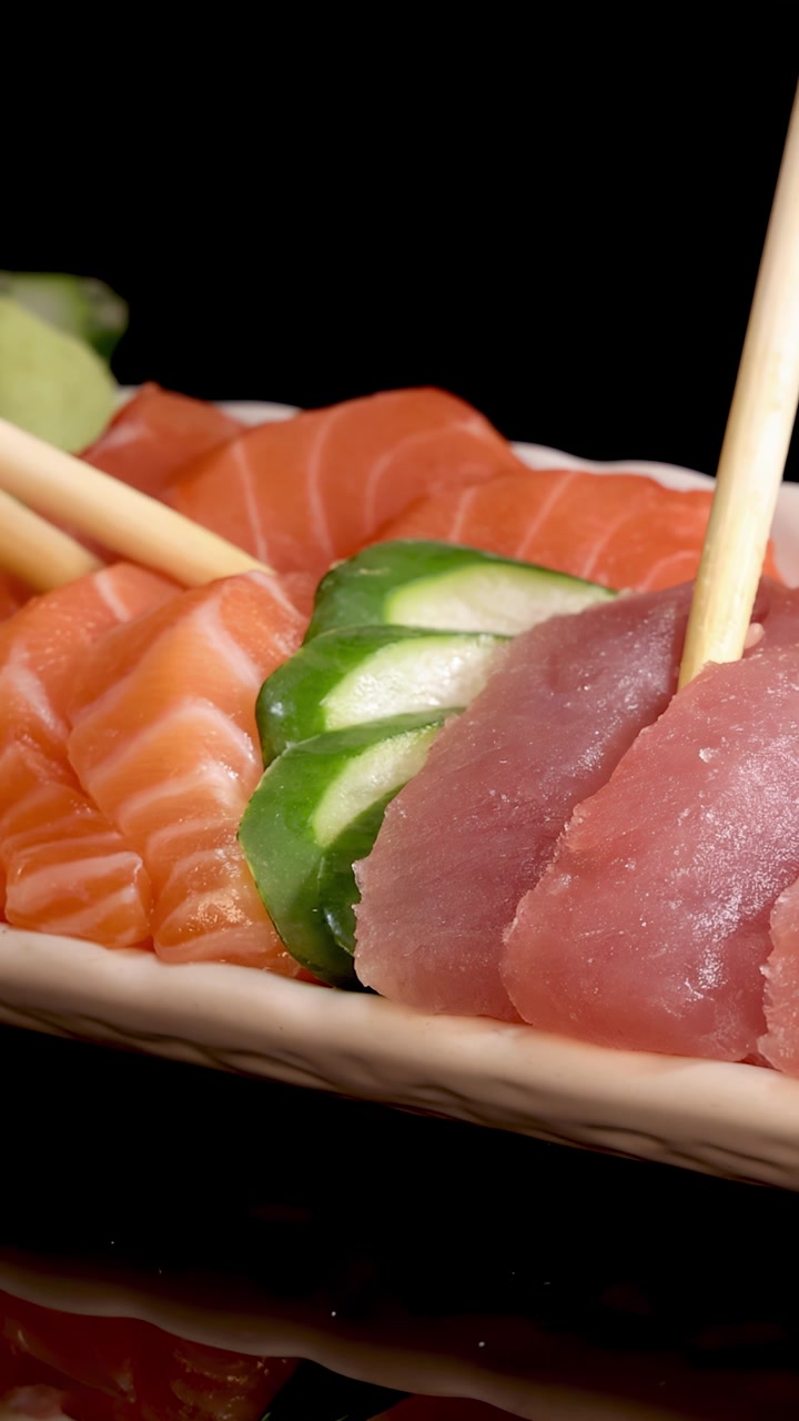 Le bacchette sollevano una fetta di sashimi di tonno fresco da un set di sushi giapponese, disposto con salmone, cetriolo e wasabi su un piatto bianco su sfondo nero. L'illuminazione da studio evidenzia i colori vivaci e le texture.