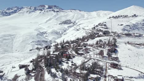 Dolly-in-aerial-view-of-the-houses-and-the-mountain-village-of-Farellones-completely-snowed-in-with-the-exclusive-resorts-of-La-Parva-and-El-Colorado,-Chile