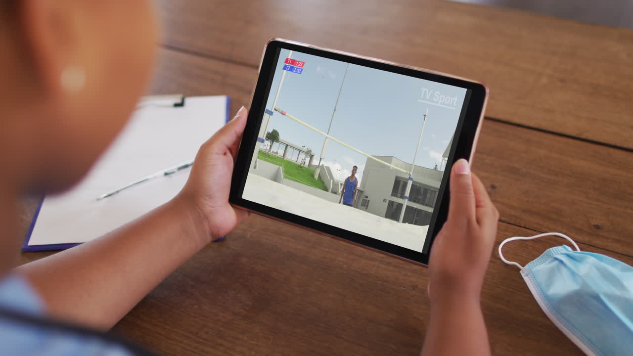 compuesto de una mujer sentada en una mesa con una máscara facial, viendo el evento de salto alto de atletismo en la tableta