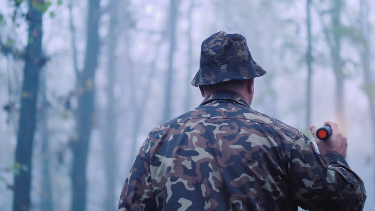 위장을 한 남자가 안개 속에서 길을 밝히는 숲 속을 걷고 있다.