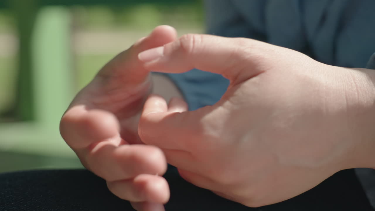 백인 여성이 손가락을 구부리고 깍지를 낀 채 무릎 위에 올려놓고 있어요. 클로즈업 핸드헬드 촬영으로 안절부절못하는 움직임을 보여주며 햇빛이 피부와 손톱을 비추고, 사색에 잠긴 긴장감은 기대감이나 초조함을 암시합니다.