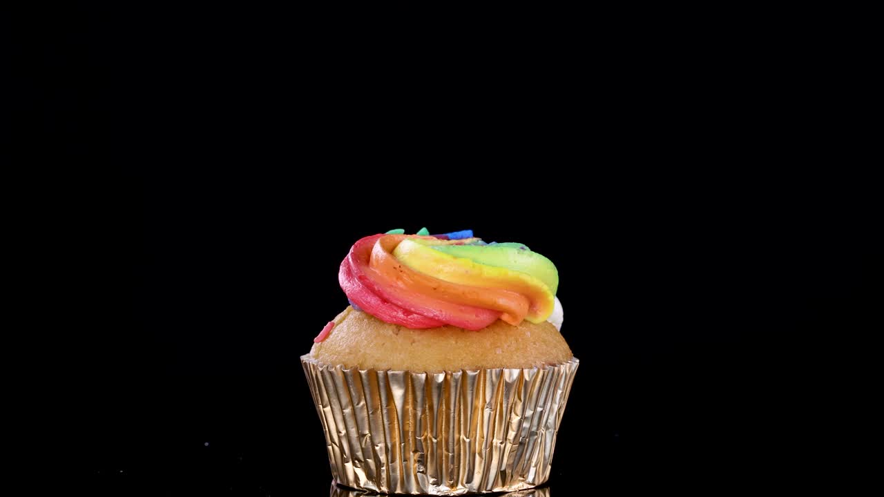 Hand places white candle into rainbow cupcake with gold wrapper on black background, minimal shadows