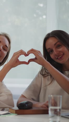 lesbian couple makes heart shape with hands expressing love and smiling. loving women sit at table in living room looking in camera closeup