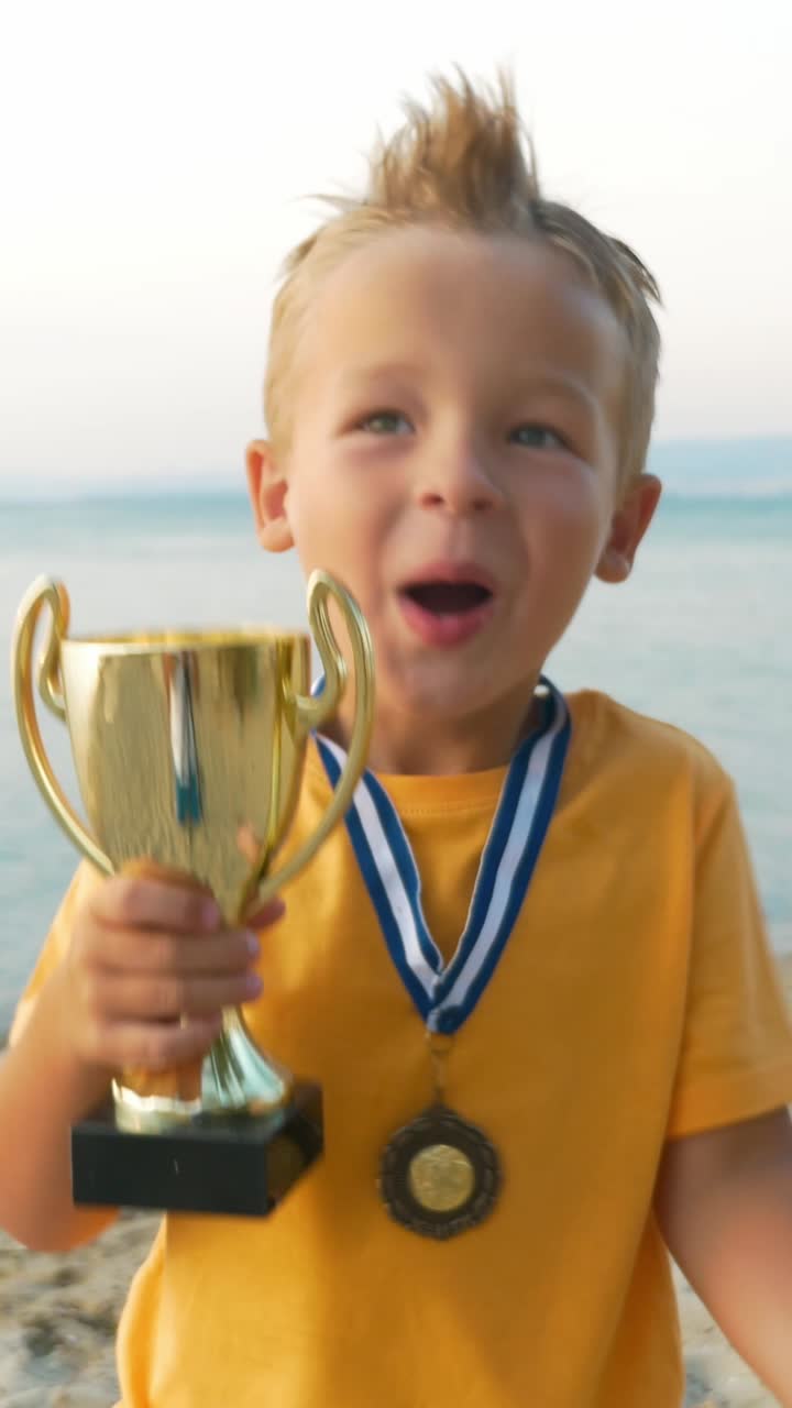 A young boy celebrating his victory at the beach with a medal and trophy