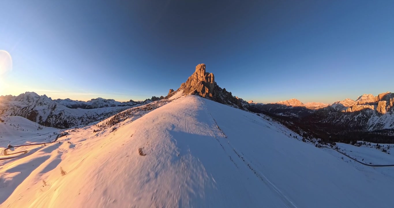 Distorted wide angle drone view of refuge with Ra Gusela rugged snow covered mountain peak illuminated by golden sunset light at Giau Pass in Italy. Aerial forward