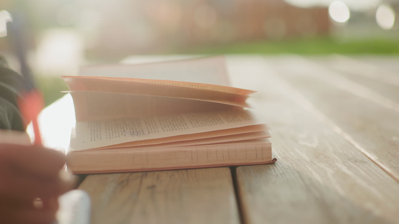 Close up of open book on wooden table outdoors with pages flipping in wind while blurred figure writes with pen in background as sunlight radiates warmly