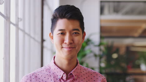millennial asian male creative standing in an office turning and smiling to camera, close up