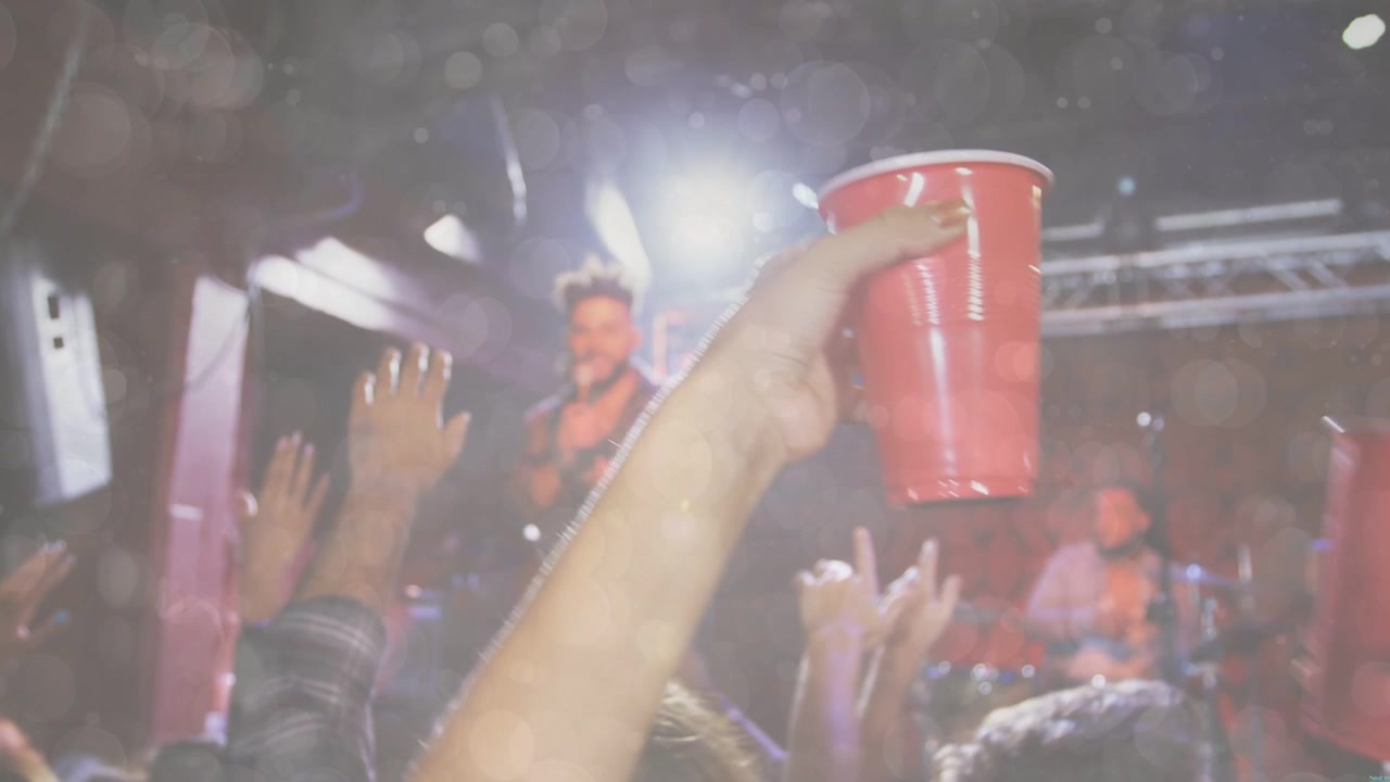 Music crowd raising two red cups while frontman singing into mic, toasting under lights and bokeh