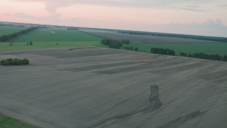 overhead drone shot capturing vast plowed field textures merging with lush green crop sections under pastel sunset sky, showcasing agriculture patterns and expansive panorama reaching distant trees