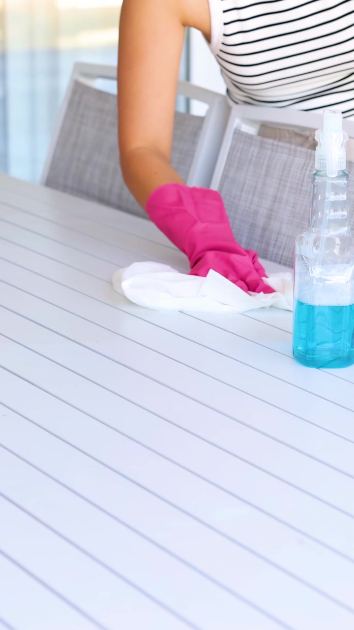 A person wearing gloves cleans a table using a spray bottle and cloth in a bright, outdoor setting
