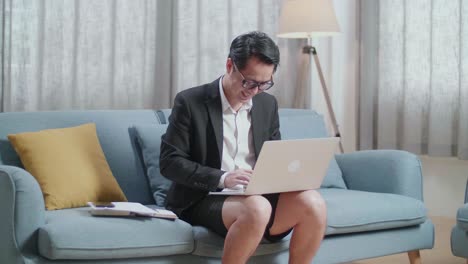 소파에 앉아서 집에서 노트북으로 타이핑하는 재과 쇼트를 입은 미소 짓는 아시아 사업가