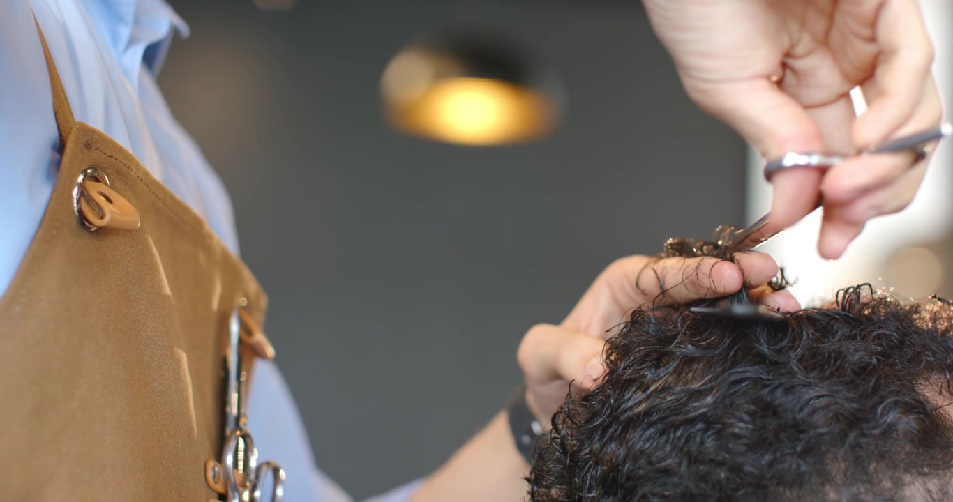 En afroamerikansk mandlig barber og en kunde i salonen, barberen i forklæde løfter krøller, klipper med saks og former håret.