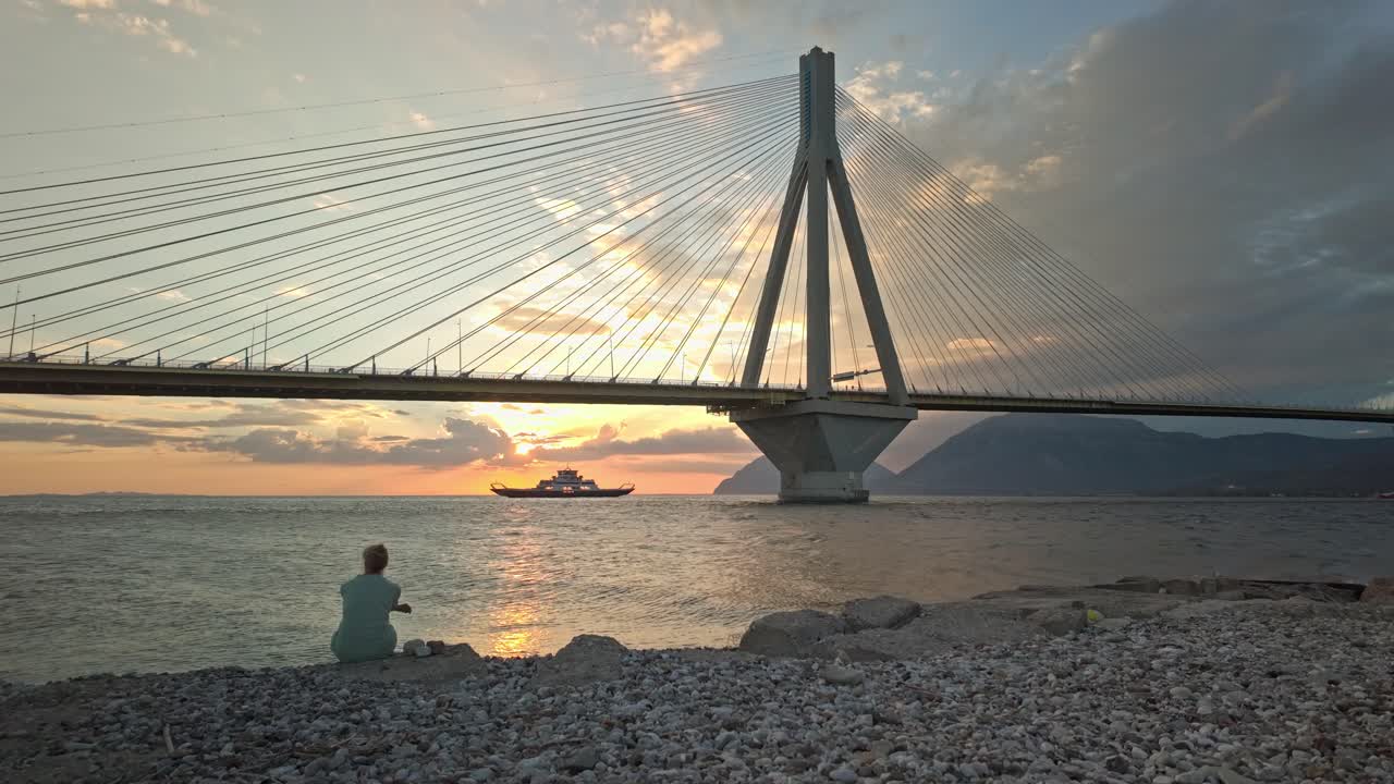 Reflective Woman sits on deserted beach watches seaferry cross Corinth bay sunset