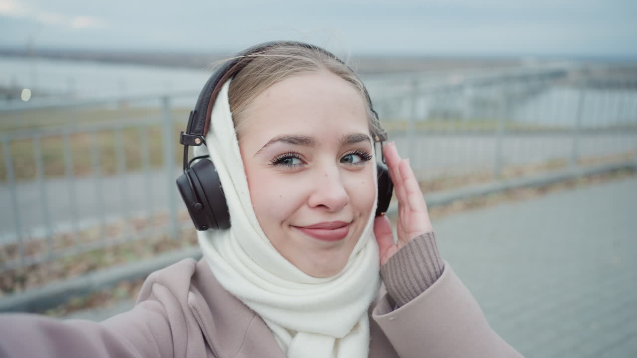 Donna che si gode la serata al fiume. Donna con sciarpa che si scatta un selfie allegro in riva all'acqua al crepuscolo. Donna rilassata con cappotto e segno della pace che cattura il momento vicino al fiume nella morbida luce serale.