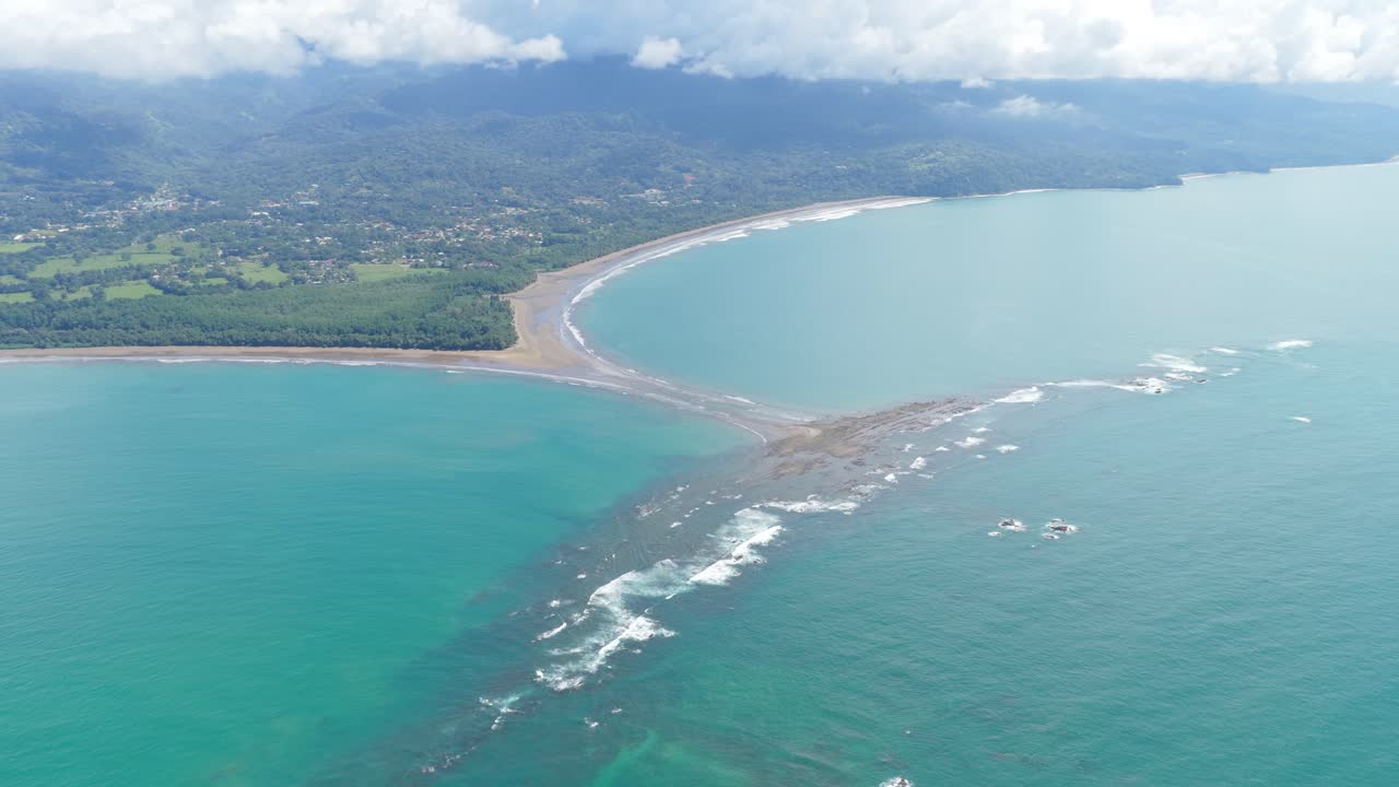 Veduta aerea di una costa tropicale con tombolo a coda di balena e onde che si infrangono.