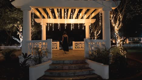 Elegant-young-woman-in-a-black-dress-walks-up-the-stairs-to-a-cosy-resort-terrace-during-an-atmospheric-night