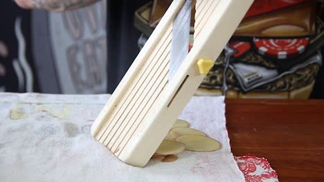 Male-tattoed-hands-slicing-raw-potato-into-thin-chips-with-a-mandolin-in-a-wooden-board-with-a-kitchen-towel-below---Slow-motion