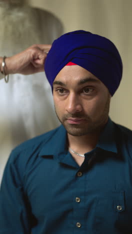 vertical video studio shot of older sikh man tying fabric for turban for younger man against plain background