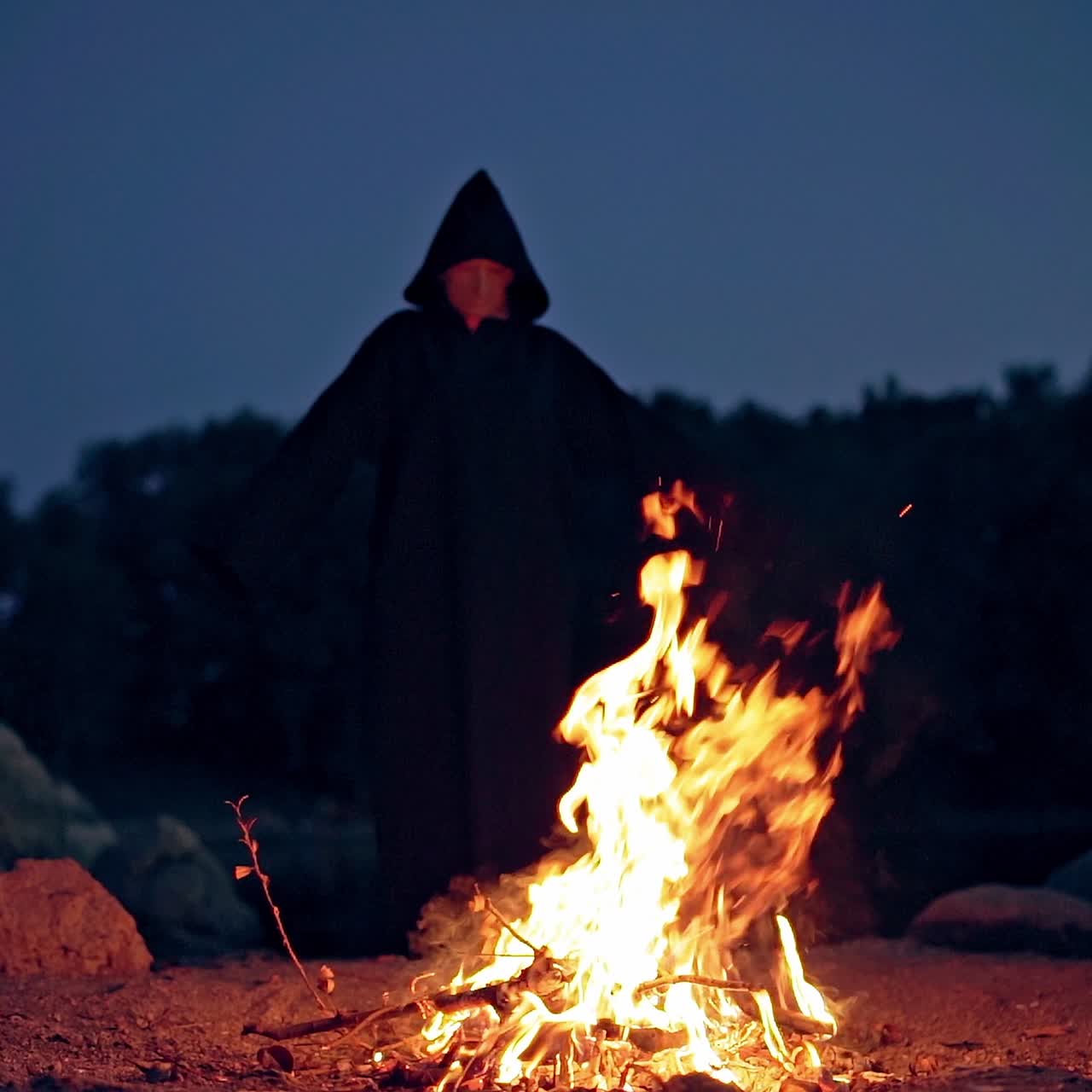 Dark wizard in hood behind the burning fire. Scary death with outstretched arms doing shamanism. Black ghost performing ritual near the fire. Horror concept.