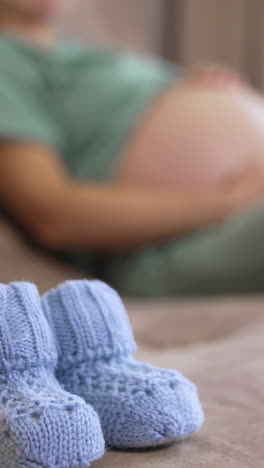 beautiful tiny knitted socks for future child. baby socks on blurred background of a living-room with a pregnant woman sitting on a couch and stroking her stomach. vertical video