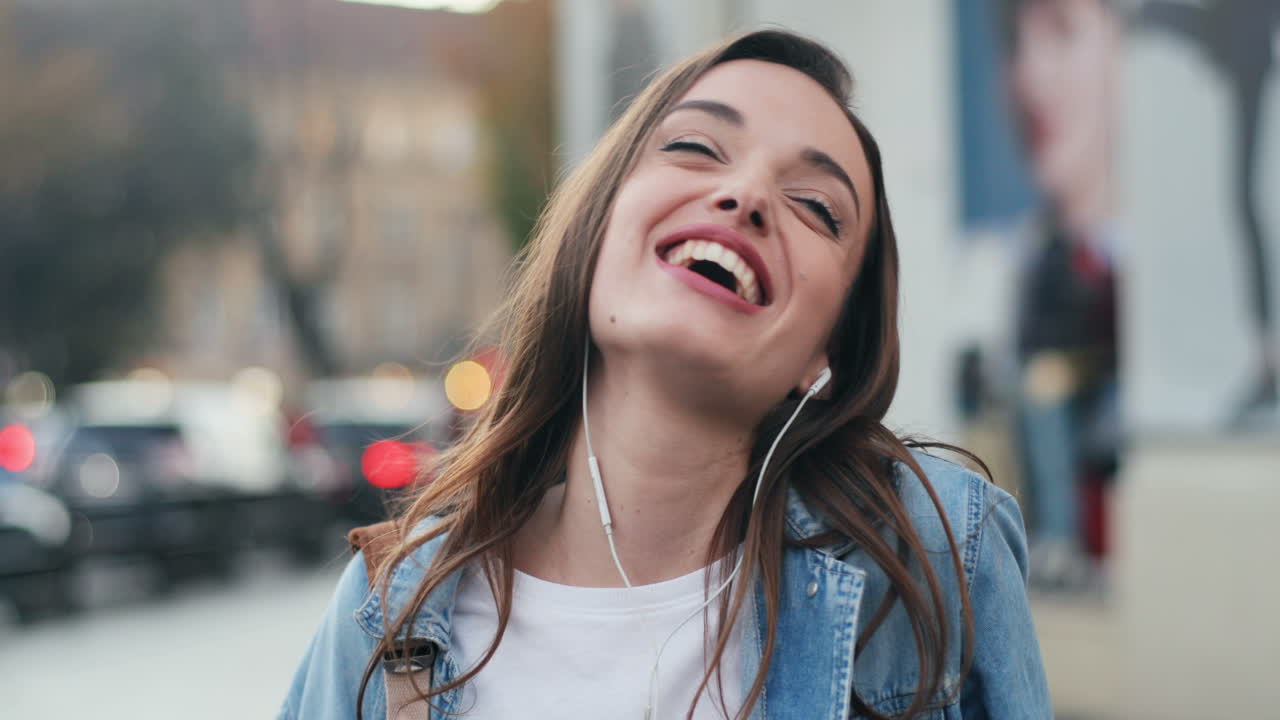 vista de cerca de una joven caucásica con auriculares escuchando música en un smartphone, cantando y bailando en la calle mientras mira la cámara