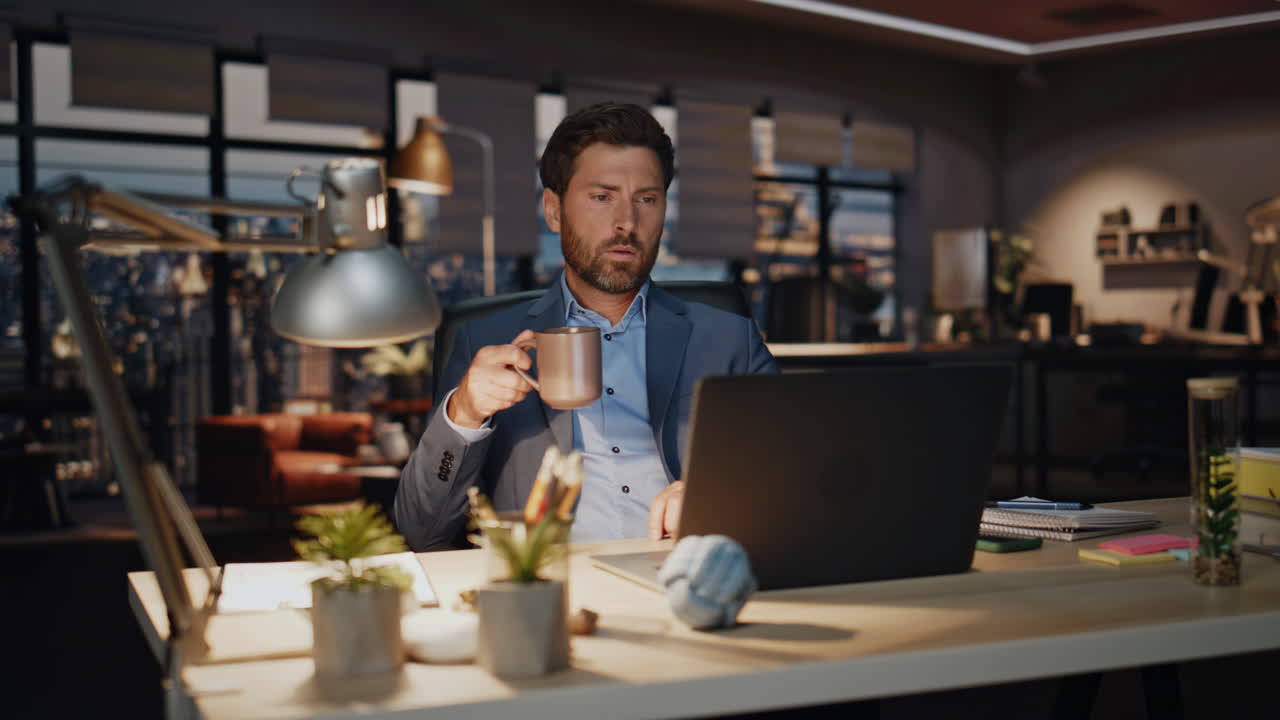 Uomo d'affari serio che guarda il computer in ufficio di notte, primo piano. Il capo beve il tè.