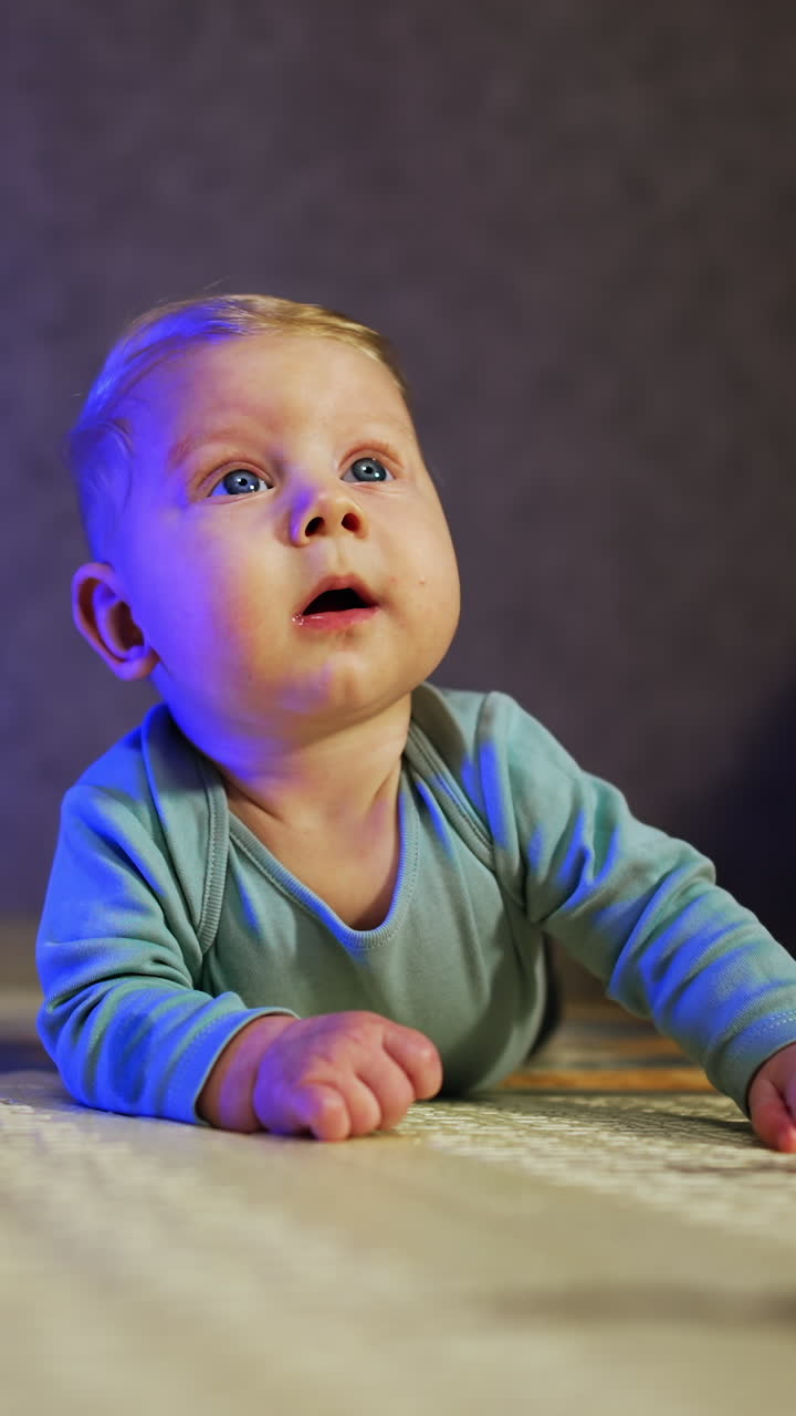 Adorable blonde baby boy lies on belly looking up with curiosity. Cute child lies at home on the floor among the toys. Vertical video