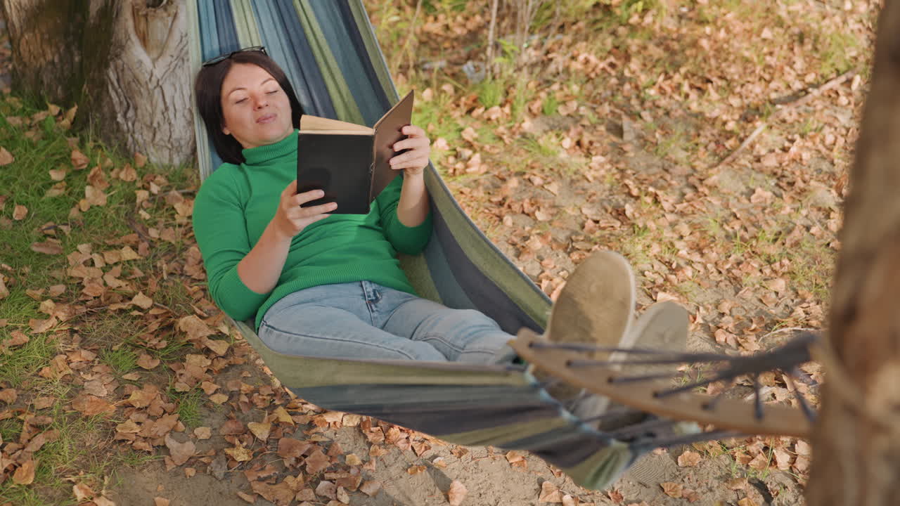 Mulher descansando na rede, cena de acampamento sob as árvores com livro e botas visíveis, vista de corpo inteiro de postura relaxada, jeans e suéter verde, chão coberto de folhas, corda e tronco de árvore, luz suave do dia.