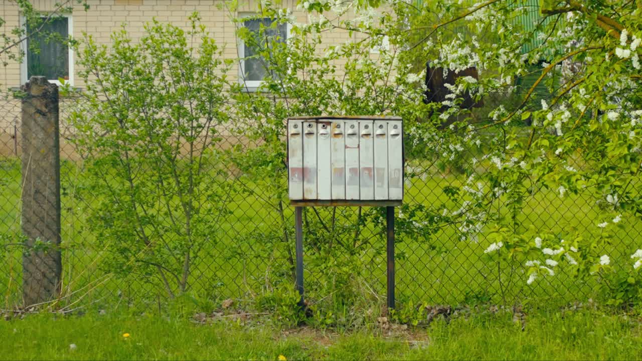 Row of Baltic weathered white metal mailboxes stands beside a chain-link fence with green grass and bushes around. Daugavpils, Latvia (Latgale)