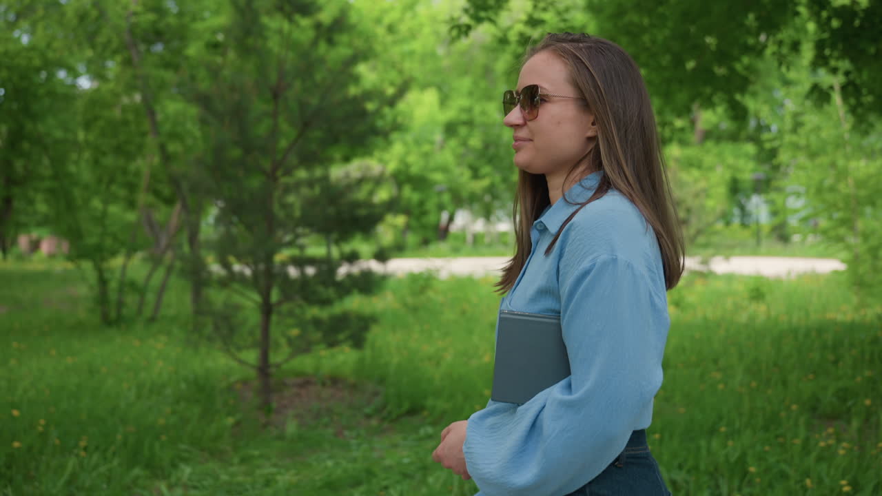 폴더를 들고, 선글라스를 착용하고, 목적 있는 걸음으로 공원을 걷는 백인 학생, 캠퍼스 분위기, 캐주얼한 블라우스와 데님, 집중된 표정, 잎이 무성한 푸른 주변 환경, 학습 통근 장면