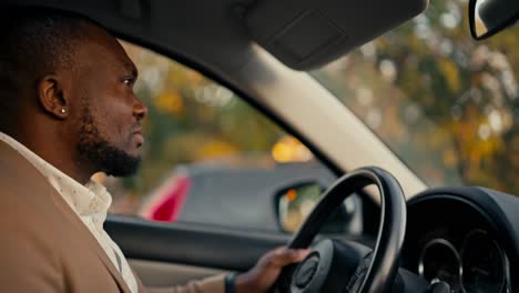 vista lateral de un hombre de negocios confiado con color de piel negra y barba en una chaqueta marrón conduce un coche durante su viaje de negocios en una ciudad moderna y mira por la ventana lateral