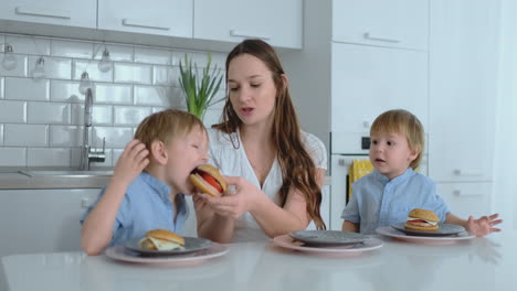 a young beautiful mother in a white dress with two children is smiling and eating fresh burgers in her kitchen. happy family homemade food healthy foods