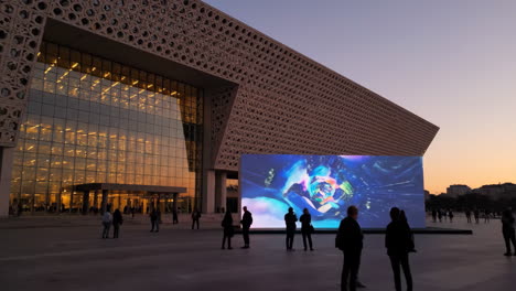 people gather to view a large outdoor digital art display on a modern building at dusk