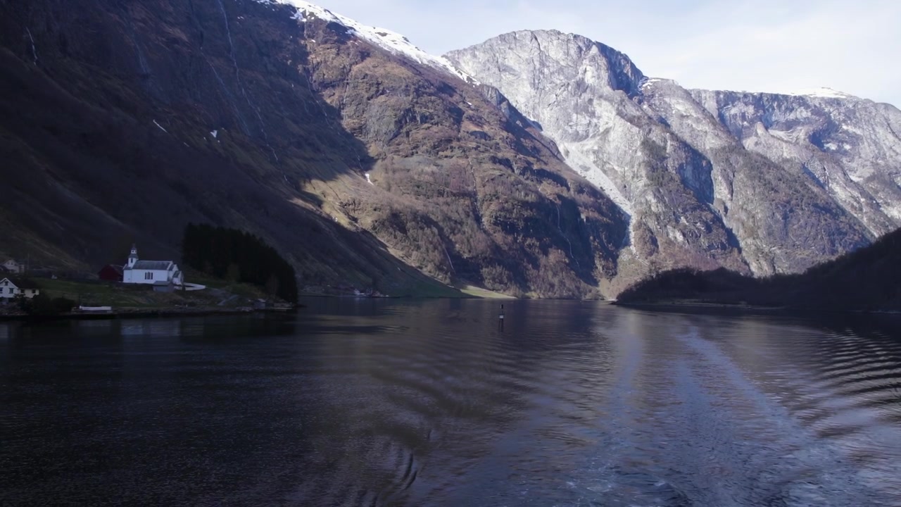 Båt beveger seg gjennom rolige norske fjordfarvann forbi en liten landsby, Bakka kirke og dramatisk fjellandskap.