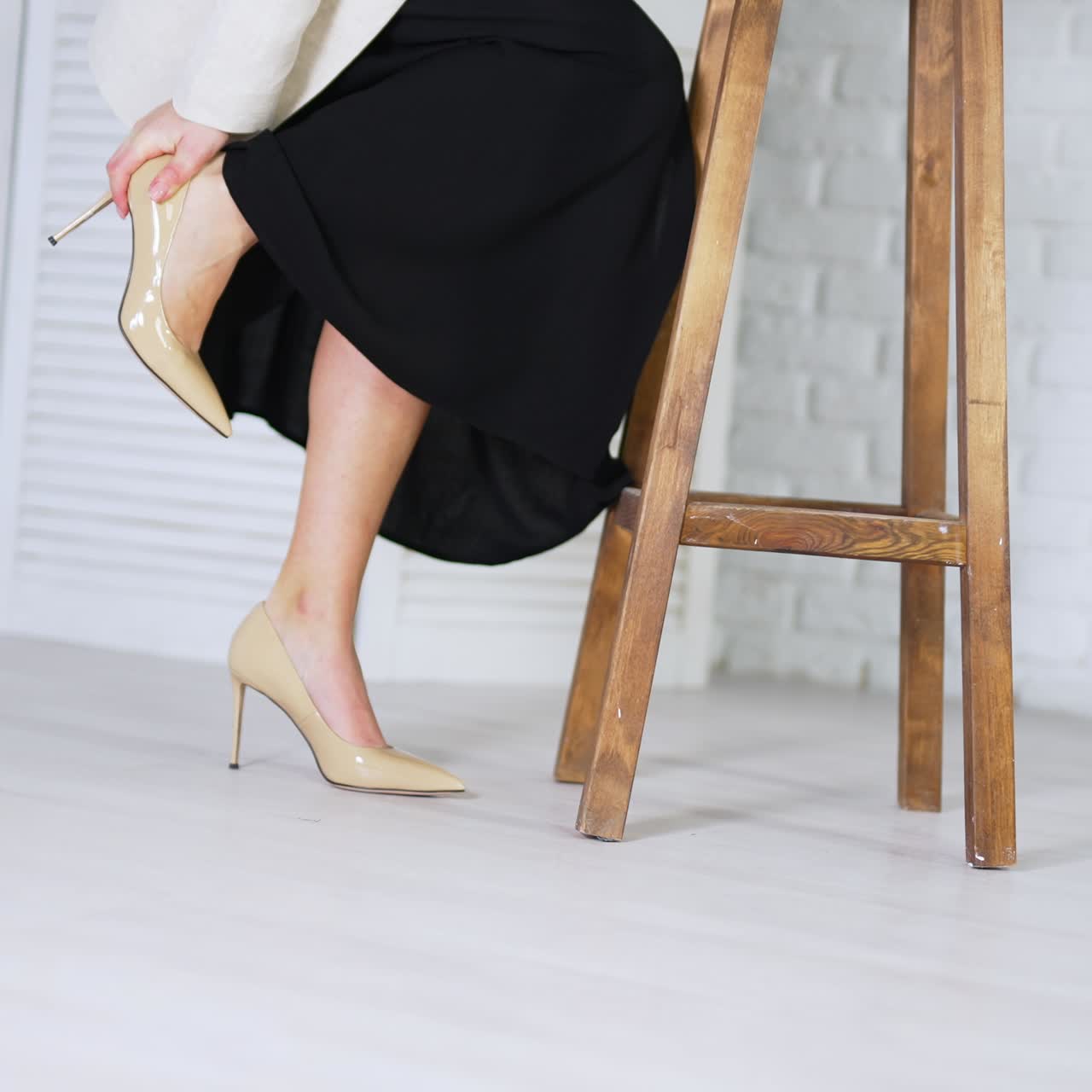 Caucasian woman in beige stiletto shoes comes up to a wooden tabouret. Model lifts her leg to feel tired foot from walking on high heels