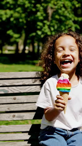 a happy young girl eating a rainbow ice cream cone on a park bench