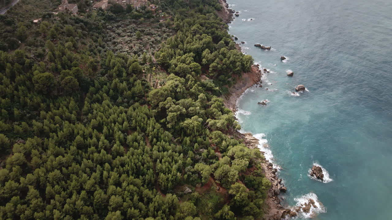Una telecamera aerea si muove in avanti, per poi inclinarsi verso l'alto partendo dalla costa di pini, rivelando un paese collinare, terreni agricoli e del fumo che si alza sulla costa della Tramuntana di Maiorca