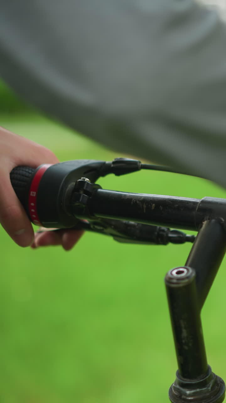회색 옷을 입은 손으로 주차된 자전거의 부분적인 시각, 드바를 잡고 조정하고, 브레이크 기능을 확인하고, 배경이 흐릿하고, 울창한 녹색 공원을 보여줍니다.