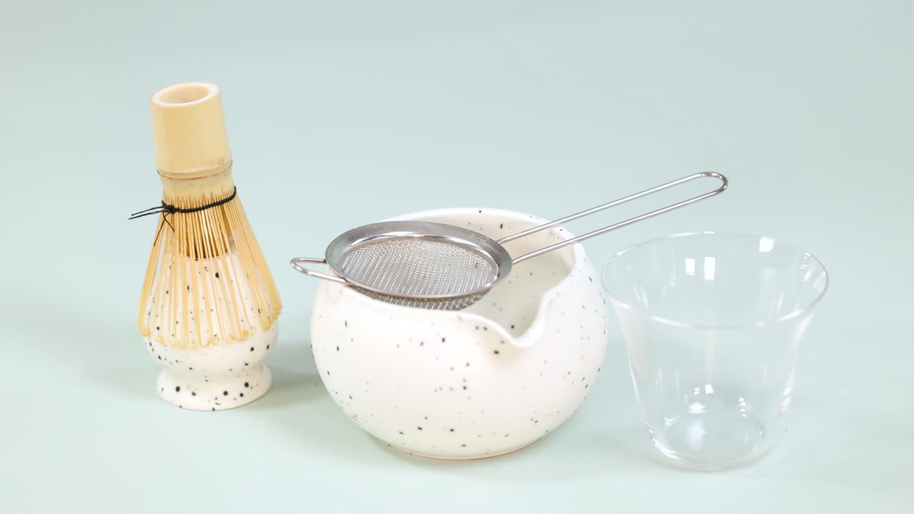 Hand sifts matcha powder through strainer into bowl, with bamboo whisk and glass cup nearby