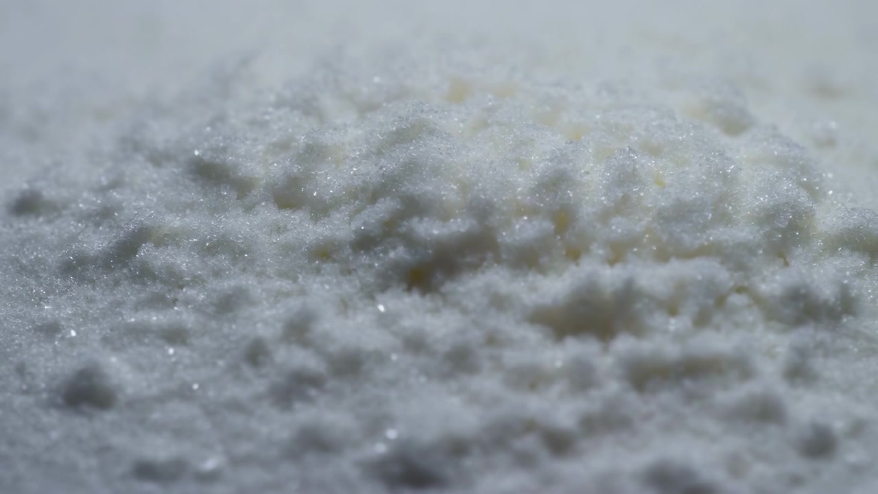 Vista orizzontale quasi macro di una superficie bianca granulare con un piccolo cumulo centrale, grani compattati e assestamento localizzato sotto una luce morbida e diffusa, che mostra una texture cristallina pallida, un leggero scintillio.
