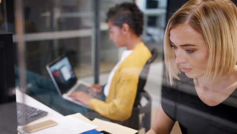 Dos-Mujeres-Jóvenes-Trabajando-Juntas-En-Computadoras-En-Un-Cubículo-De-Oficina-De-Vidrio,-Una-Volteándose-Hacia-La-Otra,-Enfocadas-En-Rack,-Vistas-A-Través-Del-Vidrio