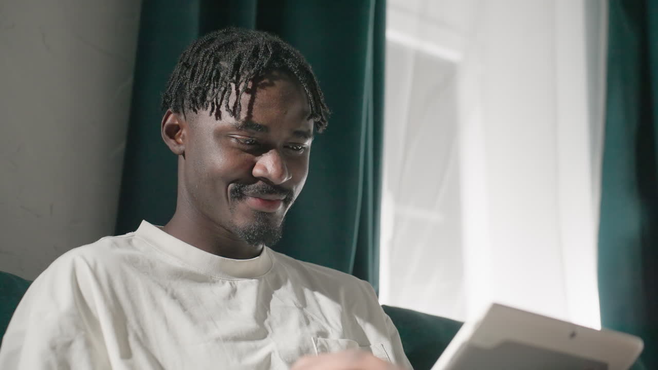 Black youth smiling while using tablet on teal sofa, relaxed and joyful. Soft natural light from window highlights his happy expression, creating a peaceful, cozy atmosphere in living room