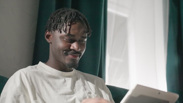 Black youth smiling while using tablet on teal sofa, relaxed and joyful. Soft natural light from window highlights his happy expression, creating a peaceful, cozy atmosphere in living room