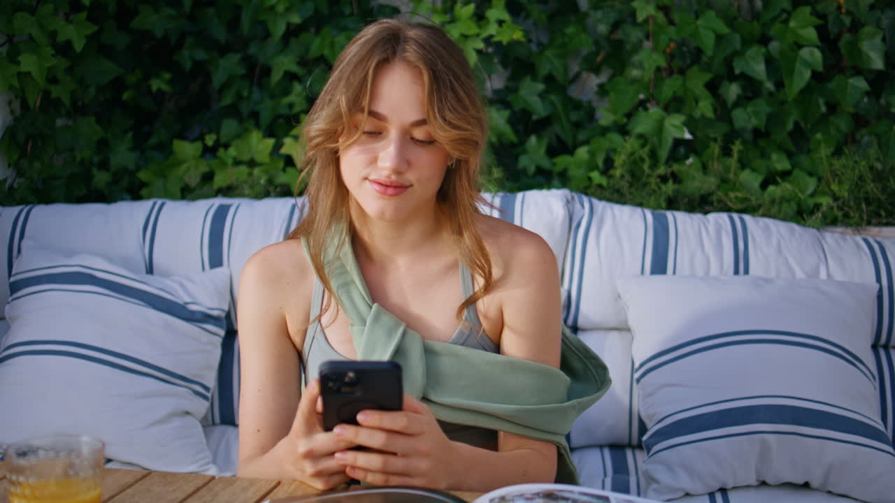 Primo piano di una donna felice che manda messaggi con il cellulare, seduta comodamente sul divano di casa in terrazza
