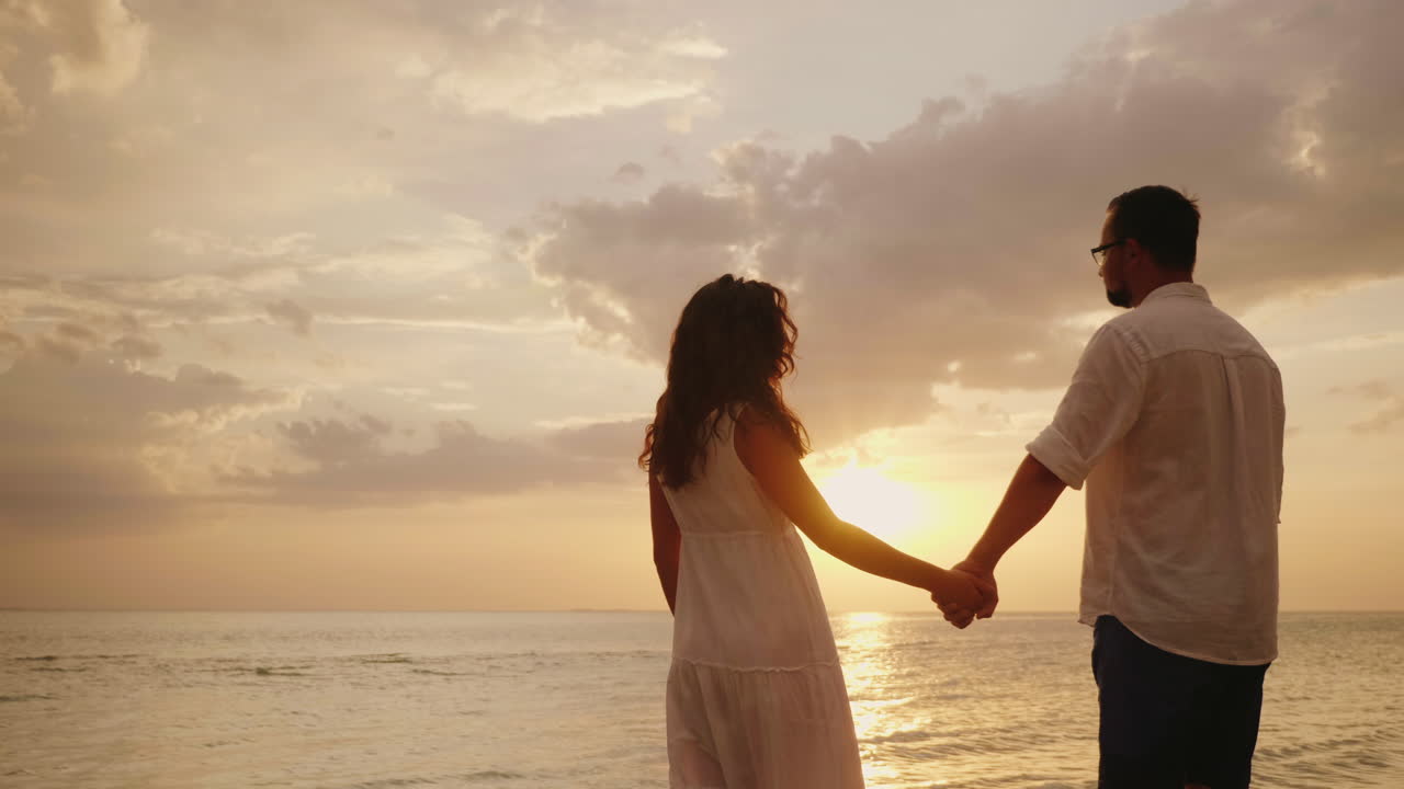 손을 잡고 있는 젊은 부부는 물 속에 서서 바다 4k 비디오 너머의 일몰을 바라본다