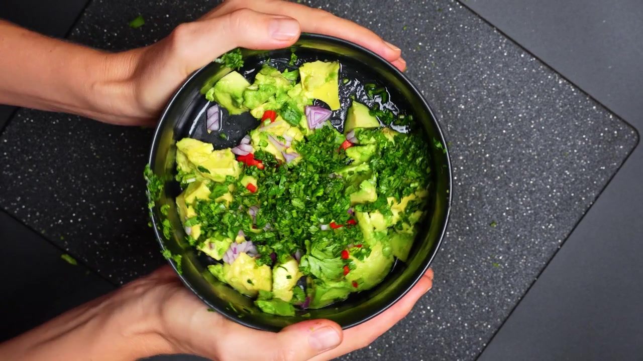 Mani che tengono una ciotola di guacamole appena fatto con coriandolo. Vista dall'alto di una persona che presenta una ciotola scura contenente avocado schiacciato, cipolle, peperoncino ed erbe tritate