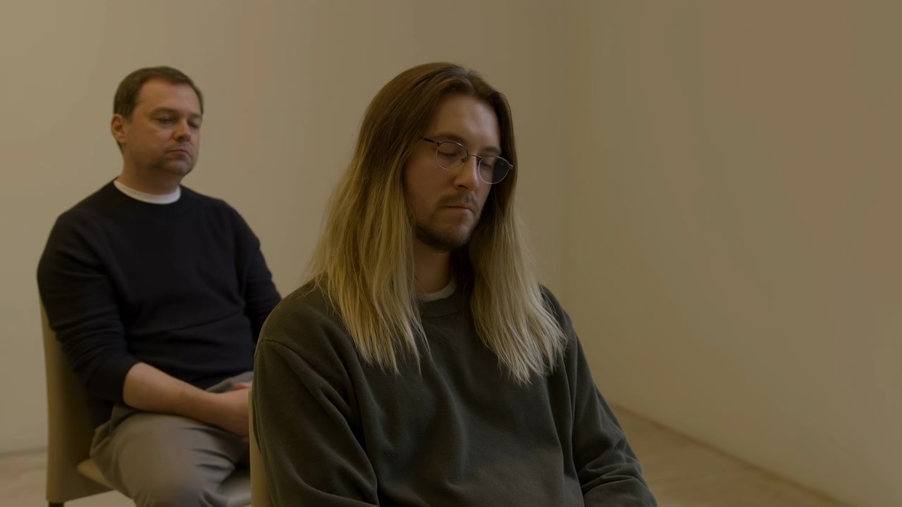 Medium shot of two men seated in a simple indoor room with a plain beige wall. Younger man with shoulder-length hair and glasses sits in the foreground